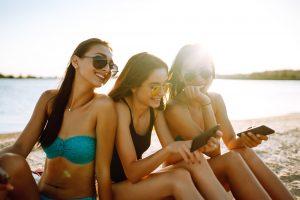 Three beautiful girls using mobile smart phone at the beach. Girls sunbathing on the beach.